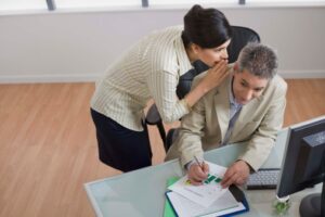 social media - A woman leans over whispering to a man at a desk. He is holding a pen, looking thoughtful. Office setting with a computer and documents on the desk.