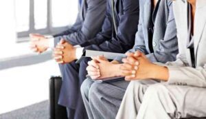 DONT BE AFRAID TO START AT THE BOTTOM Four people in professional attire sit side by side, hands clasped, conveying anticipation. The setting appears to be a waiting area, suggesting an interview.