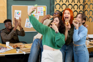 A group of five Gen Z individuals in a lively office setting enjoy pizza and takes a selfie.